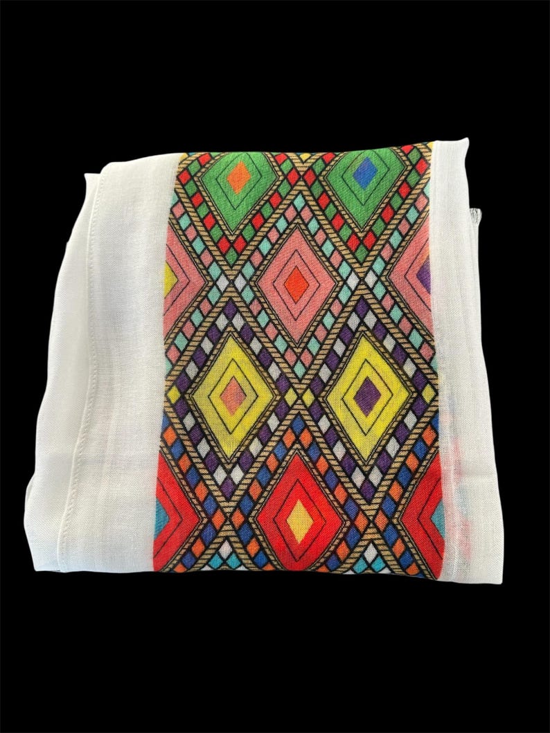 May include: White textile with a central band featuring a colorful geometric diamond pattern. The diamonds are in shades of red, yellow, green, pink, and blue, outlined with black and brown. The textile is folded and appears to be made of a lightweight fabric.