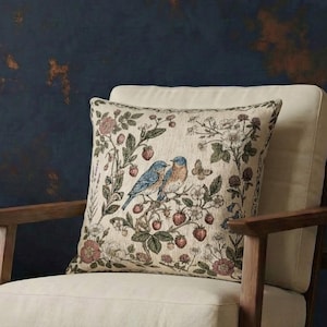 May include: A wooden armchair with a cream-colored cushion and a square throw pillow featuring a botanical print of two blue birds, strawberries, and flowers. Beside the chair sits a small wooden side table holding two teal and copper vases.