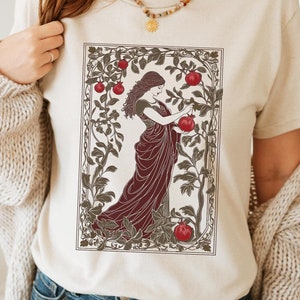 May include: A cream colored t-shirt with a graphic of a woman in a burgundy dress picking pomegranates from a tree. The tree is surrounded by a decorative frame.