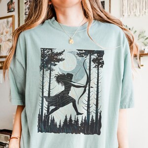 May include: A light green t-shirt featuring a black silhouette of a person with a bow and arrow, set against a forest and moon backdrop. The design is printed on a soft, comfortable t-shirt.