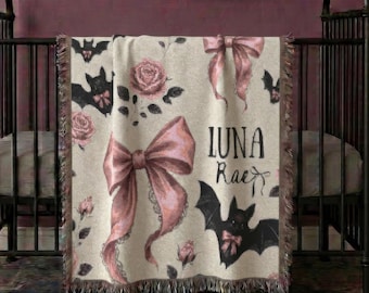 May include: A woven throw blanket featuring a repeating pattern of pink roses, dark bats with pink bows, and large pink bows on a cream background. The text Luna Rae is printed in black script. The blanket has a fringed edge and is draped over a crib.