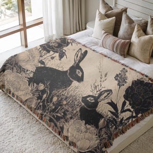May include: A beige throw blanket featuring a black and white illustration of two rabbits amidst floral and botanical elements. The blanket has a fringed edge and is draped over a bed with pillows.