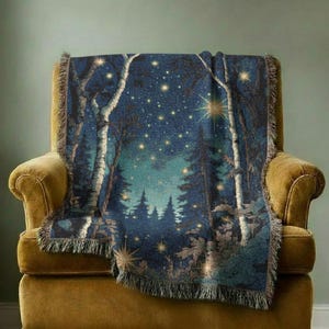 May include: A woven throw blanket featuring a nighttime forest scene with tall birch trees, dark evergreen silhouettes, and a starry sky filled with glowing yellow points of light. The blanket has a fringed edge and a deep blue and teal colour palette.