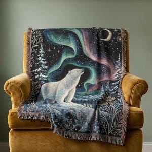 Pode incluir: Uma manta tecida apresentando um urso polar branco em uma paisagem nevada sob um céu noturno com auroras boreais vibrantes, uma lua crescente, estrelas e pinheiros, finalizada com uma borda decorativa de franjas nas laterais.
