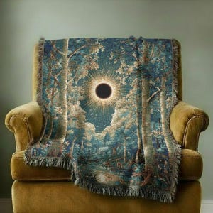 May include: A woven throw blanket featuring a detailed forest landscape with tall trees and a central solar eclipse. The design uses shades of deep blue, teal, and gold, with a fringed edge along the bottom. The scene depicts a dark sun.