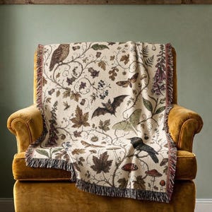 May include: A woven throw blanket featuring a cream-colored background with a detailed botanical and wildlife pattern. The design includes illustrations of an owl, a bat, a moth, a crow, various mushrooms, leaves, and flowers with fringed edges.
