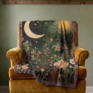 May include: A woven throw blanket featuring a dark green background with a large cream-colored crescent moon, a golden sun, and various botanical floral elements including leaves and flowers. The blanket has a fringed edge on all sides.