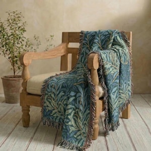 May include: A woven throw blanket featuring a repeating botanical pattern of dark blue and sage green willow leaves. The blanket has a textured finish with fringed edges and is draped over a light-toned wooden armchair with a cream-colored seat cushion.
