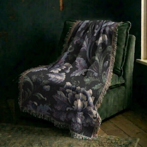 Può includere: Una coperta tessuta con uno sfondo grigio scuro e un motivo floreale su larga scala in tonalità blu tenue, verde salvia e crema. La coperta presenta un aspetto strutturato con bordi sfrangiati lungo i lati e la parte inferiore.