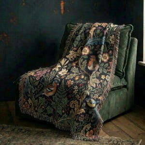 May include: A woven throw blanket featuring a dark background with a dense botanical pattern of colorful flowers, green foliage, and small birds. The blanket has a fringed edge and is draped over a dark green upholstered armchair.