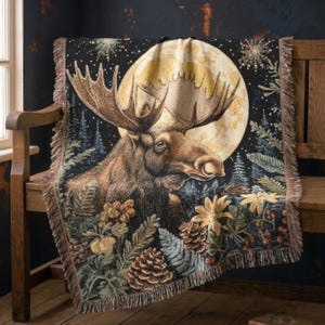 May include: A woven throw blanket featuring a detailed moose portrait centered against a large yellow full moon. The design includes dark forest trees, pinecones, ferns, and star-like accents on a black background with fringed edges.