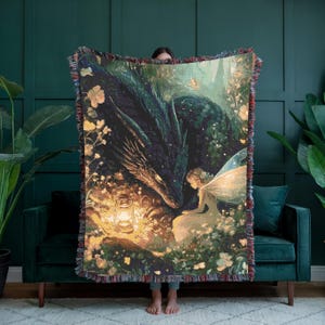 May include: A woven throw blanket featuring a fantasy scene with a dragon, fairy, and lantern. The artwork is in shades of green, gold, and blue, with a fringed edge. The dragon and fairy are the focal point of the design.