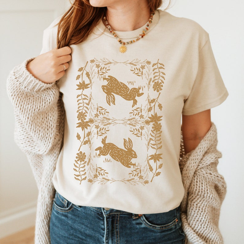 May include: Beige t-shirt featuring a gold-colored design of two leaping rabbits within a floral frame. The design includes flowers and foliage, creating a nature-inspired aesthetic. The shirt is a neutral color, suitable for various outfits.