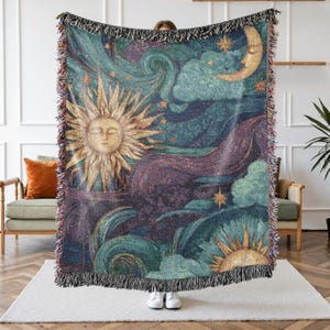 May include: Woven tapestry throw blanket featuring a celestial design with a sun, moon, stars, and clouds in shades of teal, gold, and purple. The blanket has a fringed edge and is held up by a person.