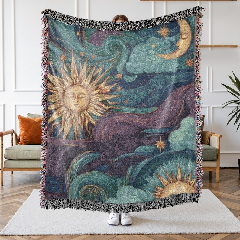 May include: Woven tapestry throw blanket featuring a celestial design with a sun, moon, stars, and clouds in shades of teal, gold, and purple. The blanket has a fringed edge and is held up by a person.