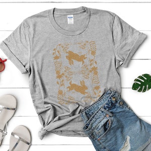 May include: A heather gray t-shirt featuring a gold-colored design of two rabbits within a floral frame. The shirt is paired with denim shorts and sandals, suggesting a casual, spring-themed outfit. The shirt is made of a soft material.