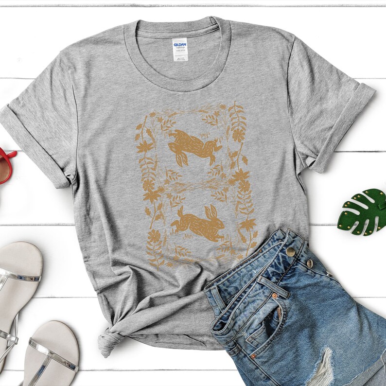 May include: A heather gray t-shirt featuring a gold-colored design of two rabbits within a floral frame. The shirt is paired with denim shorts and sandals, suggesting a casual, spring-themed outfit. The shirt is made of a soft material.