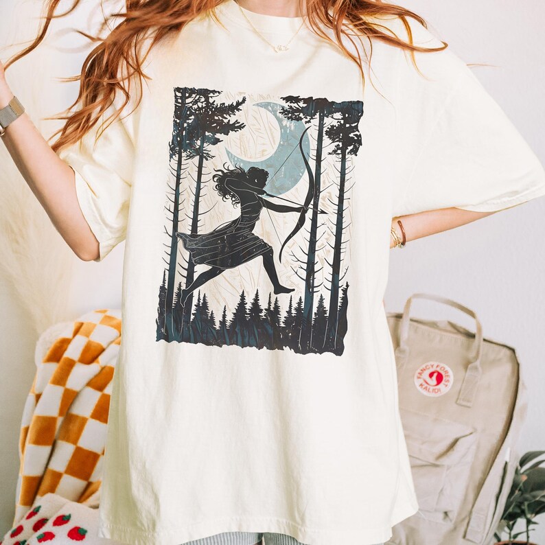 May include: Cream-colored t-shirt featuring a black and teal graphic of a figure with a bow and arrow in a forest scene with a crescent moon. The design includes tall trees and a running figure.