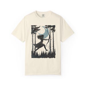 May include: Cream-colored t-shirt featuring a black and blue graphic of a person with a bow and arrow in a forest scene with a crescent moon. The design includes tall trees and a forest floor. The shirt has a crew neck and short sleeves.