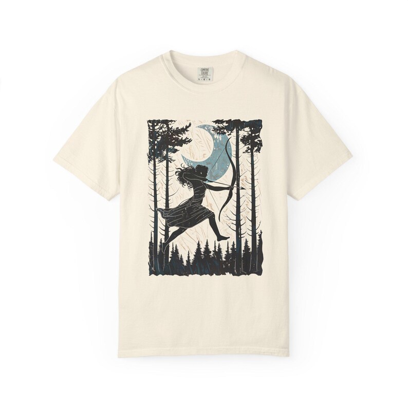 May include: Cream-colored t-shirt featuring a black and blue graphic of a person with a bow and arrow in a forest scene with a crescent moon. The design includes tall trees and a forest floor. The shirt has a crew neck and short sleeves.