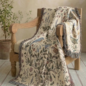 Op de afbeelding: Een beige sprei met een botanisch en vogelontwerp, gedrapeerd over een houten stoel. Het patroon van de deken bevat blauwe, groene en bruine tinten. Het heeft een gefranjerde rand.