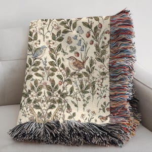 Op de afbeelding: Een geweven plaid met een cr&egrave;mekleurige achtergrond en een gedetailleerd botanisch patroon van groene bladeren, kleine bloemen, rode bessen en diverse vogels. De deken is afgewerkt met lange, veelkleurige franjes langs de randen.