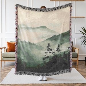 May include: A woven throw blanket featuring a misty mountain landscape in shades of green and beige. The design includes trees and rolling hills, with a fringed edge in multiple colors. The blanket is held up by a person.