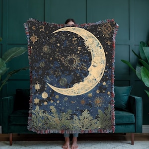 May include: A woven throw blanket featuring a large golden crescent moon with a human face, surrounded by golden stars and celestial patterns against a dark blue and black background, with a fringe border and green foliage at the bottom edge.