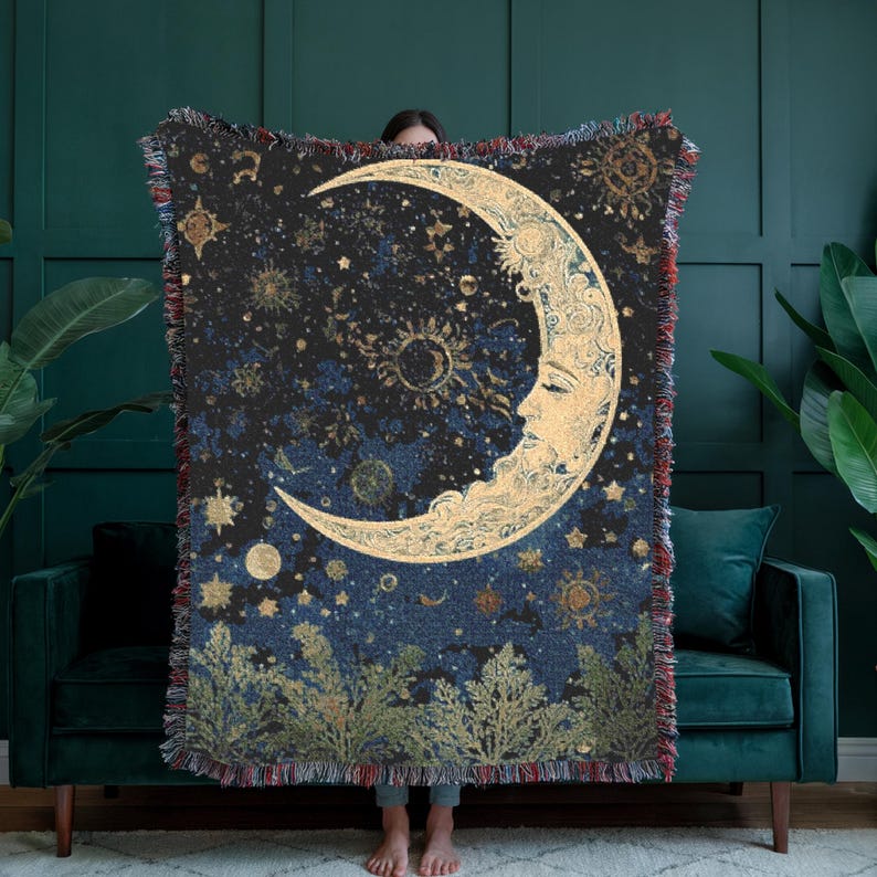 May include: A woven throw blanket featuring a large golden crescent moon with a human face, surrounded by golden stars and celestial patterns against a dark blue and black background, with a fringe border and green foliage at the bottom edge.