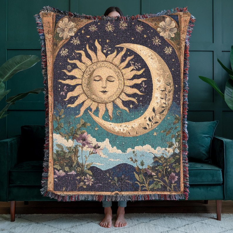Puede incluir: Una manta tejida que presenta un dise&ntilde;o central de sol y luna creciente en tonos dorados y crema sobre un fondo azul oscuro. La secci&oacute;n inferior muestra un paisaje con flores y nubes, acabado con un borde decorativo de flecos.