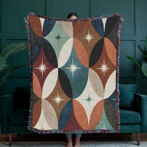 Può includere: Una coperta tessuta con un motivo ripetitivo di grandi ovali sovrapposti in tonalità arancio ruggine, verde acqua, crema e nero. Il design è accentuato da piccole stelle bianche a più punte centrate all'interno delle forme ovali.