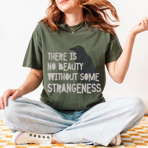 May include: A person wearing a green t-shirt with a black crow graphic and the text "There is no beauty without some strangeness". The person is sitting with their legs crossed and wearing blue jeans.