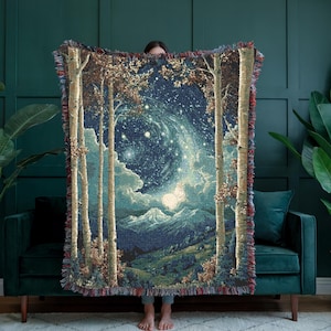 Puede incluir: Manta tejida con un diseño de escena de bosque con abedules que enmarcan un cielo estrellado. La manta tiene un borde con flecos con detalles en rojo, marrón y azul. El diseño incluye montañas, nubes y una galaxia en espiral.