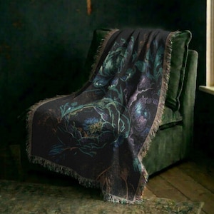 May include: A dark-colored woven throw blanket featuring a large-scale floral pattern in shades of teal, blue, and purple. The blanket has a textured surface with visible fringe detailing along the edges, draped over a dark green upholstered chair.