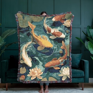 May include: Woven throw blanket with a koi pond design. The blanket depicts koi fish in orange, yellow, and white in a teal pond with lily pads and flowers. The edges have a fringed trim.