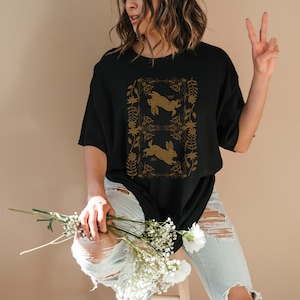 May include: Black t-shirt featuring a gold-colored design of two rabbits within a floral frame. The rabbits are depicted in a running pose. The shirt is a casual style, with a relaxed fit.