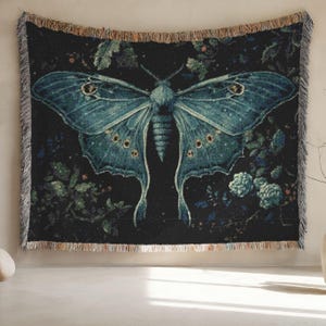 May include: A woven tapestry featuring a large, detailed moth with teal wings against a dark background. The moth has intricate patterns and is surrounded by floral elements. The tapestry has a fringed edge.