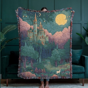 May include: A woven throw blanket featuring a detailed tapestry design of a tall castle set against a dark green sky with a large yellow moon and stars. The scene includes dense forest foliage, pinkish clouds, and a small white deer in the foreground.