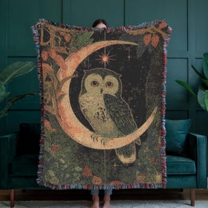 May include: A woven throw blanket featuring an owl perched on a crescent moon against a dark background. The design includes stars, foliage, and a color palette of black, brown, green, and orange. The blanket has a fringed edge.