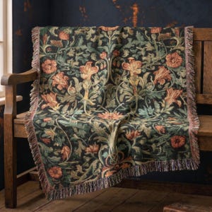 May include: A woven throw blanket featuring a dark black background with a repeating botanical pattern of orange flowers and green foliage. The rectangular textile has a decorative fringed edge along the sides and is draped over a wooden bench.