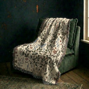 May include: A cream-colored woven throw blanket featuring a detailed botanical pattern with green leaves, red berries, blue flowers, and small brown birds. The blanket has a fringed edge and is draped over a dark green upholstered armchair.