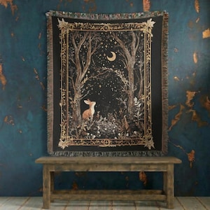 Op de afbeelding: Een zwarte sprei met een bosscène met een konijn, bomen en een halve maan. De sprei heeft een decoratieve rand en franjes. Het kleurenpalet omvat zwart, bruin en goud.
