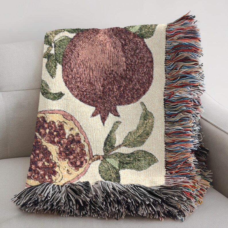 May include: A woven throw blanket featuring a cream-colored background with a large dark brown pomegranate and a halved pomegranate showing red seeds. The design includes green leaves and stems, finished with a multi-colored fringed edge.
