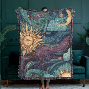 May include: A woven tapestry blanket featuring a celestial design with a sun, moon, stars, and swirling clouds in shades of gold, teal, purple, and blue. The blanket has a fringed edge.