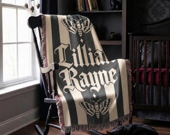 May include: A woven throw blanket featuring a black and cream vertical striped pattern, two moth illustrations with skull markings, and the name Lillia Rayne printed in a large, gothic-style font. The blanket has a fringed edge finish.