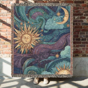 May include: Woven throw blanket featuring a celestial design with a sun, moon, stars, and clouds in shades of gold, teal, purple, and blue. The blanket has a fringed edge and is displayed against a brick wall.