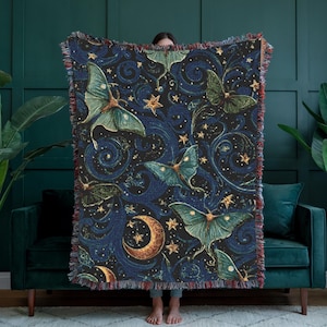 May include: A dark blue throw blanket with a celestial design featuring moths, stars, and crescent moons. The moths are teal and green, and the stars and moons are gold. The blanket has a fringed edge.