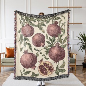 May include: A woven throw blanket featuring a cream-colored background with a repeating pattern of dark red pomegranates and green leaves. The design includes whole pomegranates and one halved pomegranate, finished with a multi-colored fringe edge.