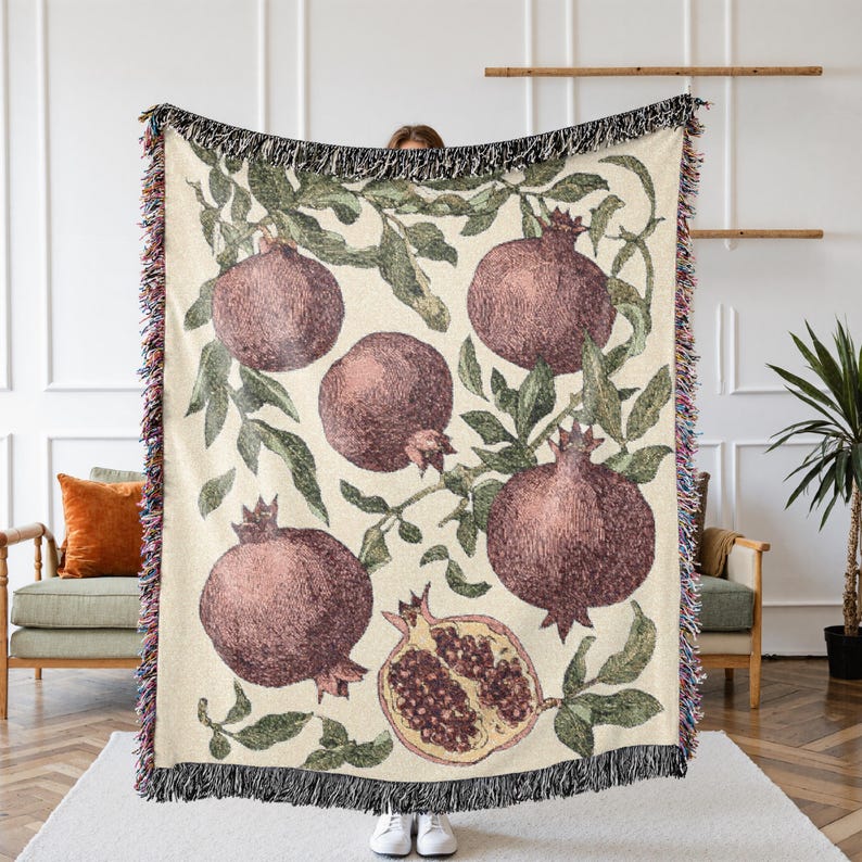 May include: A woven throw blanket featuring a cream-colored background with a repeating pattern of dark red pomegranates and green leaves. The design includes whole pomegranates and one halved pomegranate, finished with a multi-colored fringe edge.