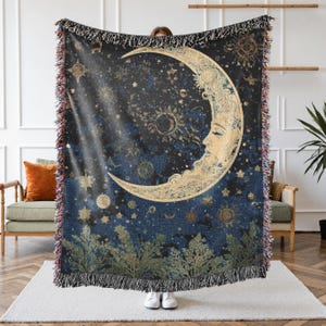 May include: A woven throw blanket featuring a dark blue night sky with a large, golden crescent moon with a human face. The design includes scattered stars, smaller celestial bodies, and green foliage at the bottom edge with fringed borders.